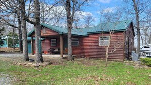 A rustic log cabin with a green roof, a porch with outdoor seating, and a small yard with trees and a gravel path. - Year Around View Lake Tenkiller (Vian)