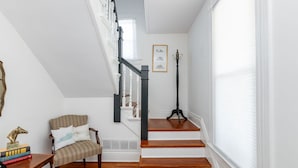 Una escalera con barandillas blancas y escalones de madera, una silla con cojines estampados y una escultura de metal decorativa sobre una mesita.