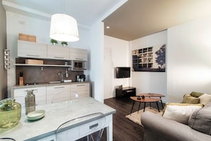 Een moderne keuken met een ingebouwde magnetron, een eethoek met een tafel en stoelen, en een woonkamer met een bank en een wandgemonteerde flatscreen-tv.
