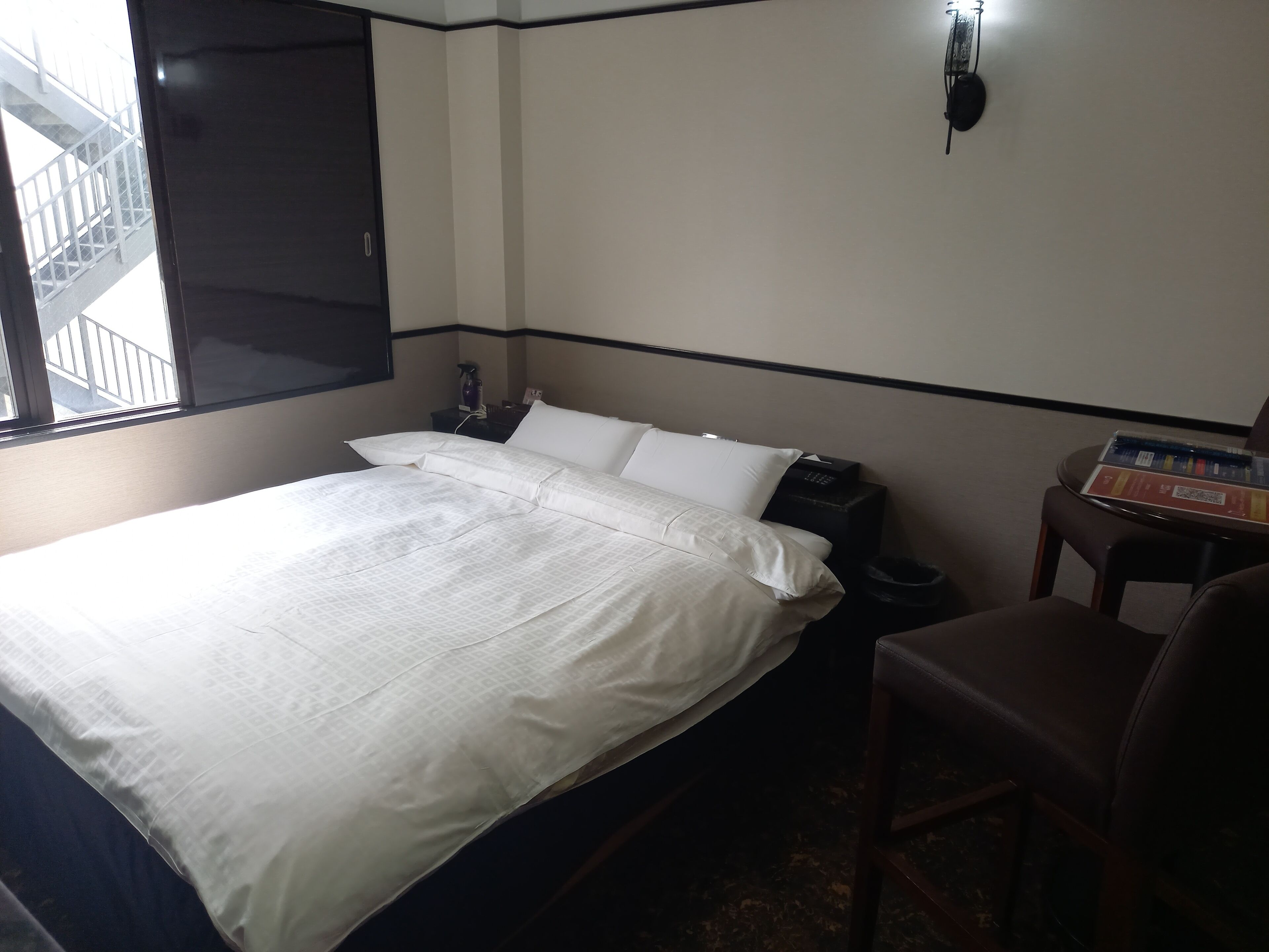 A hotel room with a bed, a nightstand, a chair, and a window with a view of an external staircase.