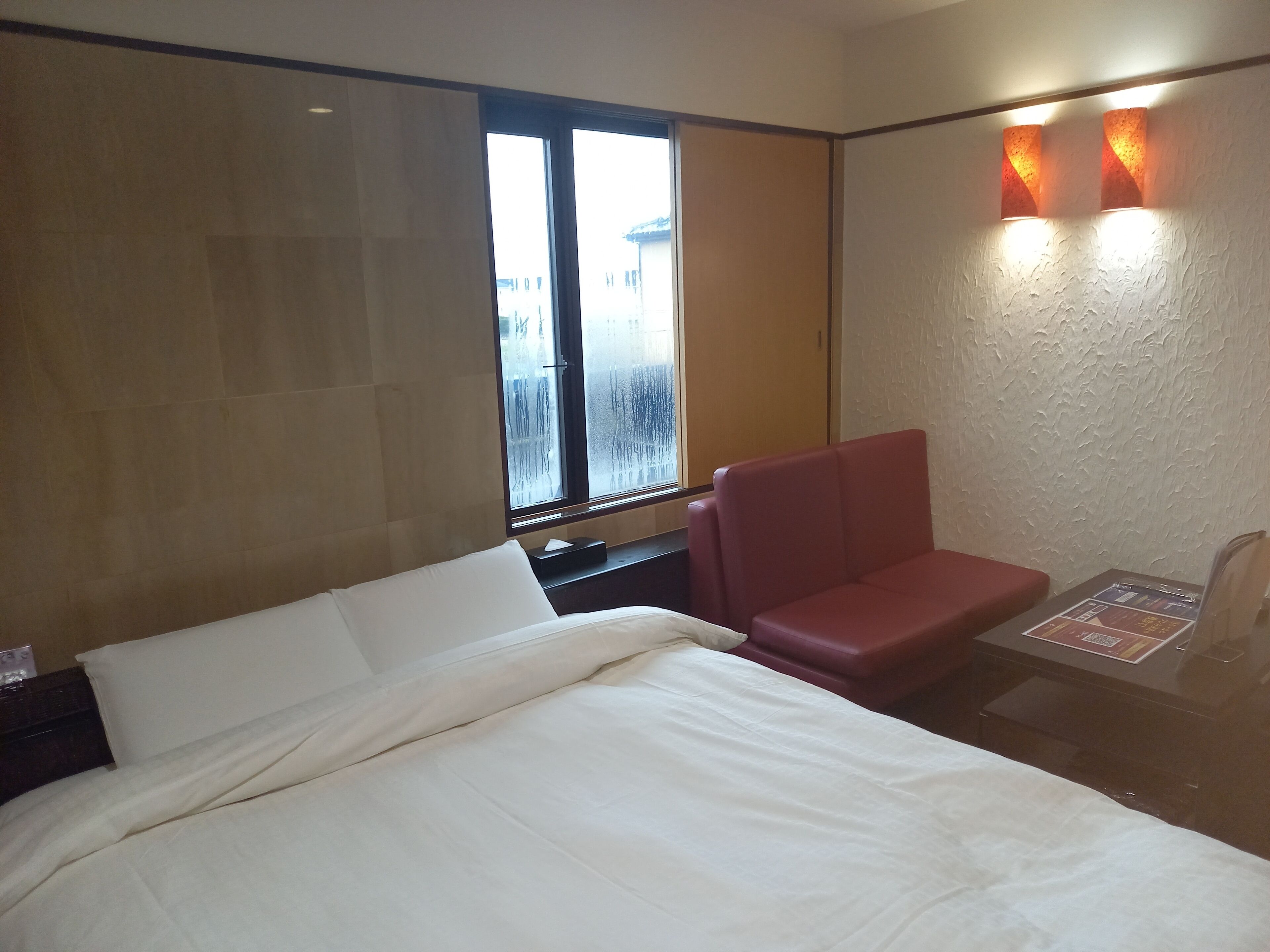 Een hotelkamer met een bed, een rode stoel, een klein tafeltje met een menukaart en een raam met geëtste glas.