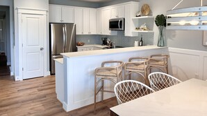 A modern kitchen with white cabinetry, a stainless steel refrigerator, a microwave, and a dining area with wicker chairs and a white table. - "Nomadica"-Modern, Top Floor Beachfront Amazing Views & Beach Access (Galveston)