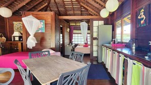 Une pièce avec un plafond en bois, une table à manger avec des chaises, un lit recouvert d’une couverture rose, une petite cuisine avec un évier et une cuisinière, et une fenêtre avec des rideaux.