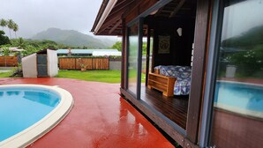 Un espace piscine avec une terrasse en bois, une pièce fermée par des baies vitrées et dotée d’un lit, et une vue sur une chaîne de montagnes.