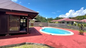 A pool area with a red surface, a building with a red roof, and a wooden fence.