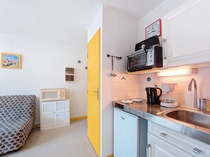 Uma cozinha compacta com micro-ondas, torradeira, cafeteira e pia. Há uma cama com cobertura estampada e um armário pequeno.