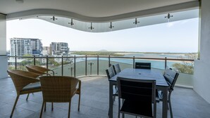 A balcony with a view of the sea and buildings, furnished with wicker chairs and a table.