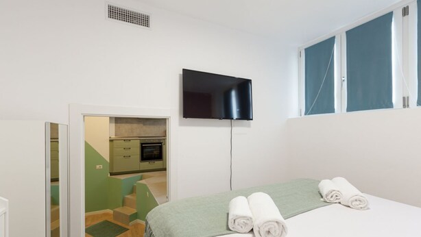 A hotel room with a bed, a flat-screen TV mounted on the wall, a window with curtains, and a kitchenette in the background. - GuestHost - Insubria Park - Pastel Colored Apartment! (Milano)