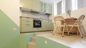 A kitchen with light green cabinets, a built-in oven, and a dining area with a table and chairs. - GuestHost - Insubria Park - Pastel Colored Apartment! (Milano)