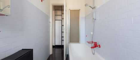 A bathroom with white tiled walls, a white bathtub, a black trash bin, and a white rug.