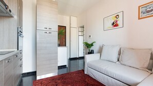 A modern living room with a sofa, a red rug, a kitchen area with appliances, and a hallway leading to another room. - GuestHost - Corvetto M3 - Comfortable Apt. with balcony! (Milano)