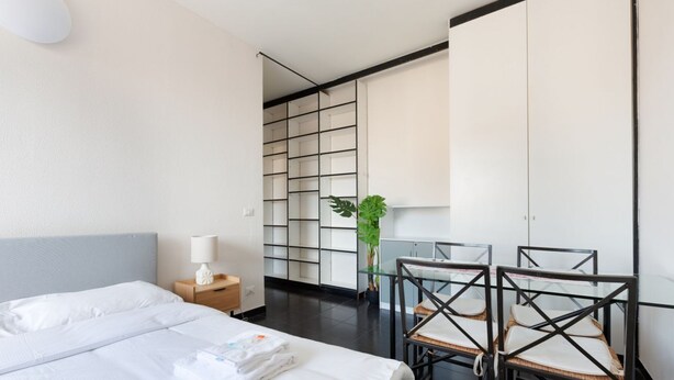 A modern hotel room with a bed, bedside table, lamp, chair, and a plant. - GuestHost - Corvetto M3 - Comfortable Apt. with balcony! (Milano)