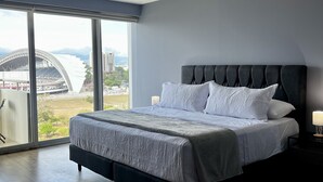 Quarto moderno com uma cama grande, uma mesa de cabeceira e vista para um estádio através de grandes janelas.