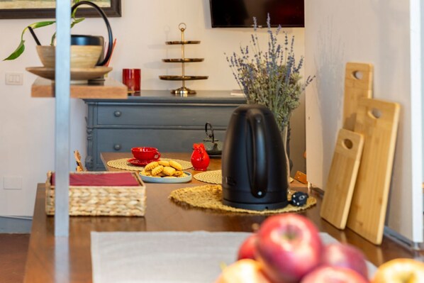 Une cuisine équipée d’une machine à café, d’une planche à découper, d’un saladier rempli de fruits et d’un vase de fleurs.
