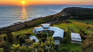 Una casa moderna con techo curvo, rodeada de exuberante follaje y vistas al océano al atardecer.