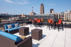 Terraza en la azotea con mobiliario exterior, incluyendo un sofá azul y sillas de mimbre, con vistas a un paisaje urbano con edificios modernos.