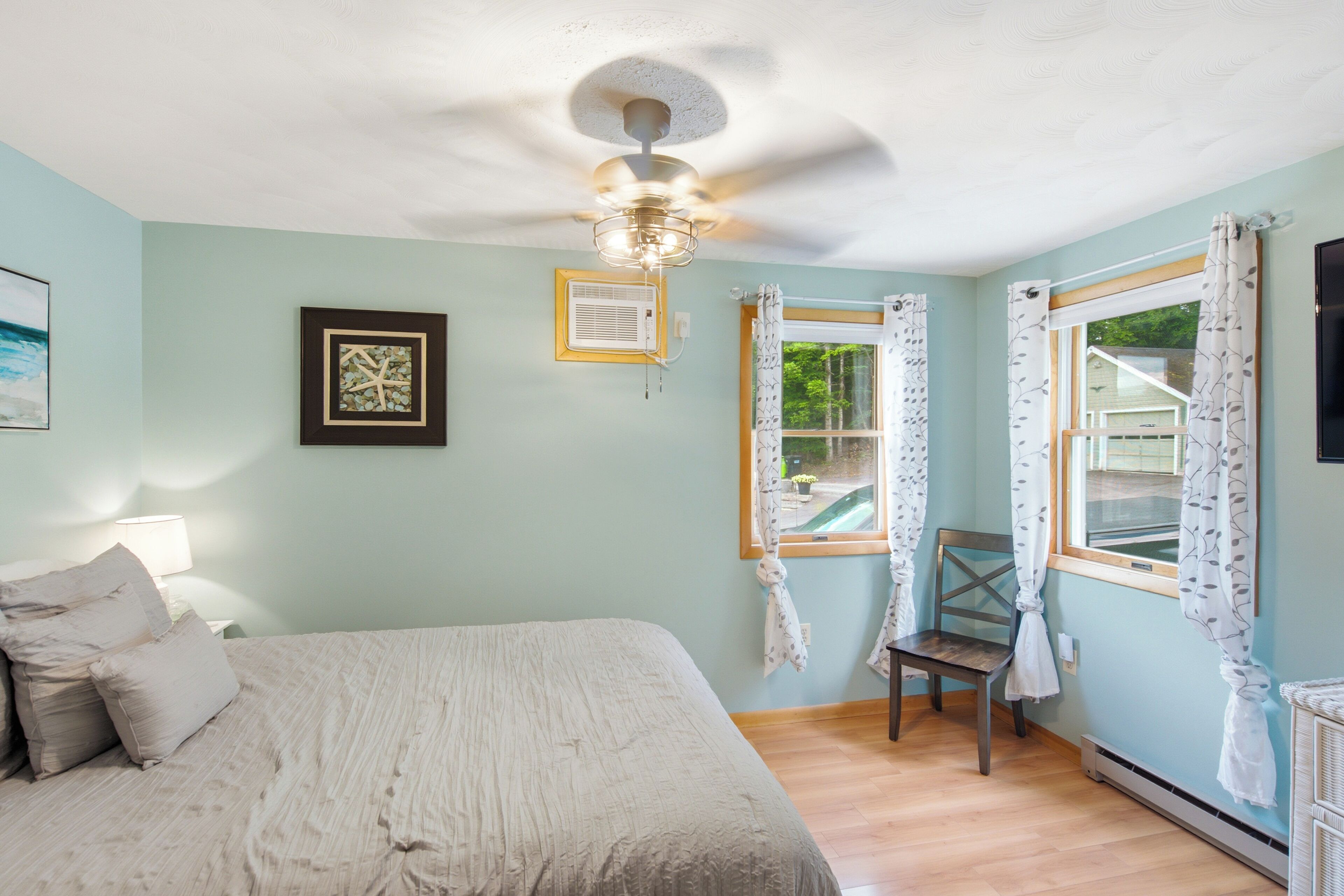 A bedroom with a bed, a chair, a ceiling fan, a window with curtains, and a wall-mounted air conditioner.