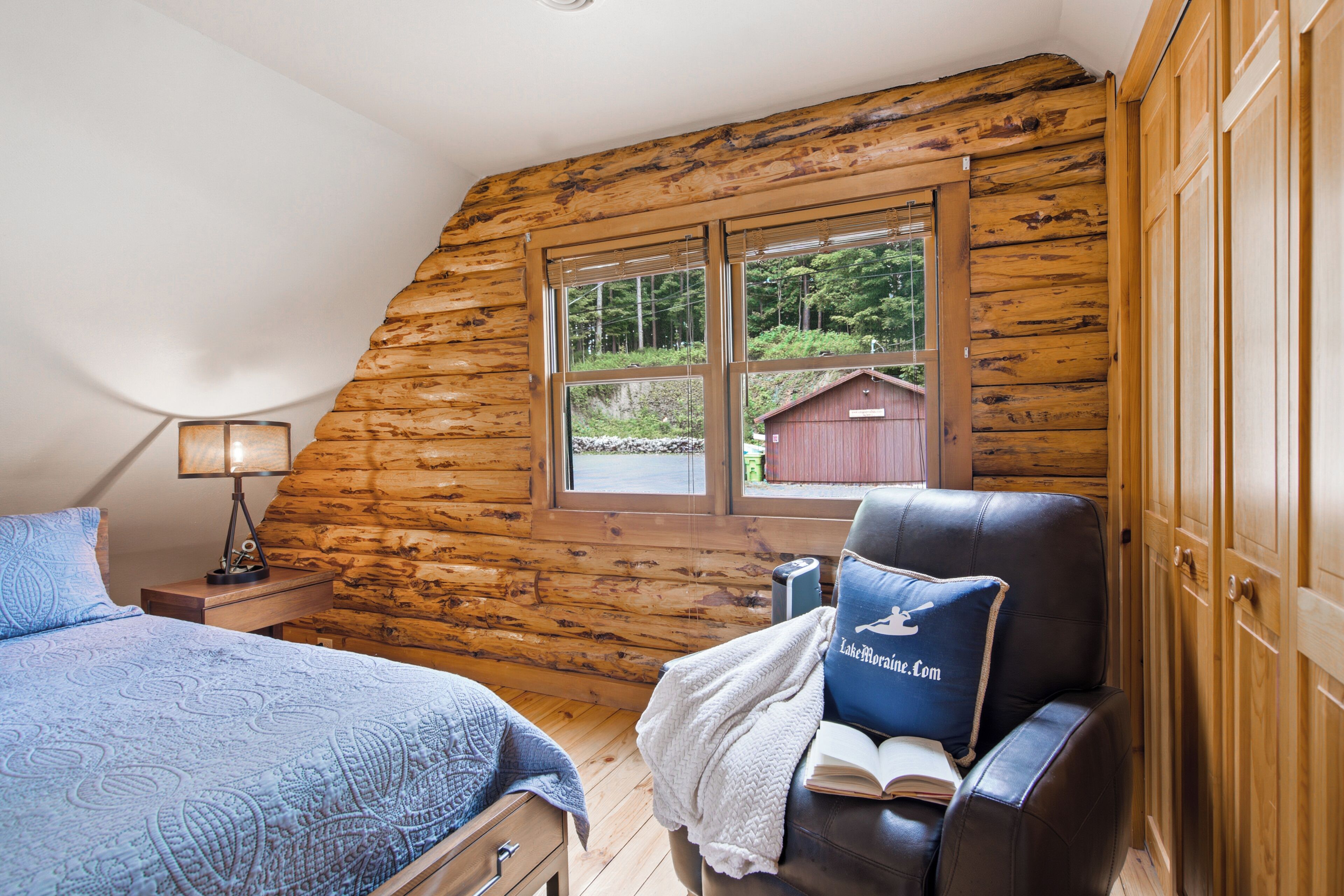 Quarto de cabana de tronco com uma cama, uma cadeira, uma mesa de cabeceira, um abajur e uma janela com vista para um prédio vermelho e árvores.