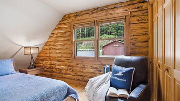Quarto de cabana de tronco com uma cama, uma cadeira, uma mesa de cabeceira, um abajur e uma janela com vista para um prédio vermelho e árvores.
