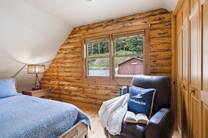 Ein Schlafzimmer in einer Blockhütte mit Bett, Stuhl, Nachttisch, Lampe und einem Fenster mit Blick auf ein rotes Gebäude und Bäume.