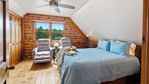 Une chambre avec un plafond en pente, des murs en bois, un lit avec une literie bleue, une chaise, une petite table avec une lampe et une fenêtre donnant sur un paysage verdoyant.