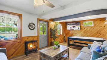 Um quarto acolhedor com piso de madeira, ventilador de teto, dardos, mesa de bilhar e lareira.