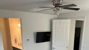 Een slaapkamer met een bed, een plafondventilator, een aan de muur bevestigde televisie en een kleine tafel met een plant.