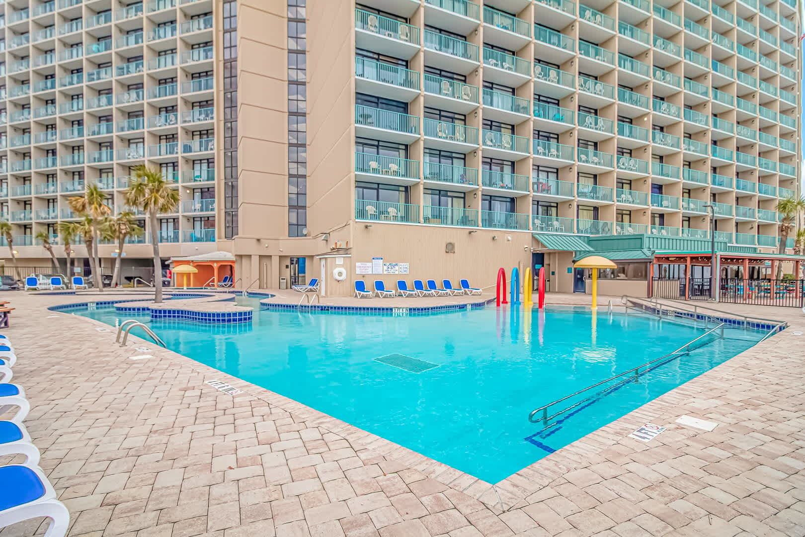 A hotel pool area with a clear blue pool, blue lounge chairs, and a large building with multiple balconies in the background.