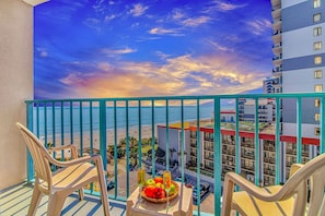 A balcony with chairs, a table with fruit and drinks, and a view of the beach and ocean.
