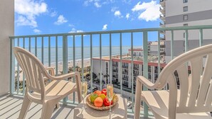 A balcony with a view of the beach, two chairs, a table with fruit and a glass of champagne, and a railing.
