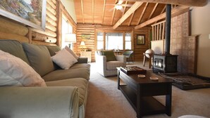 Interior acolhedor de uma cabana de madeira com sofá, poltronas, uma mesa com xícaras de chá, lareira e um quadro na parede.