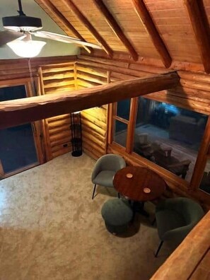 A log cabin room with a high ceiling, a ceiling fan, a large window, and a small round table with two chairs.