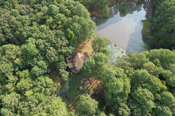 Vue aérienne d’une maison entourée d’une forêt dense et d’un étang.