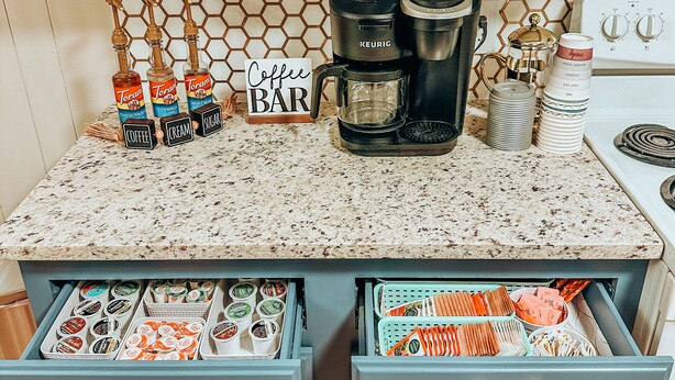 Een keuken met een koffiehoek, inclusief een koffiezetapparaat, Torani siropen en een bordje met de tekst 'Coffee Bar'. Er zijn lades met diverse etenswaren en een betegelde achterwand met een honingraatpatroon.
