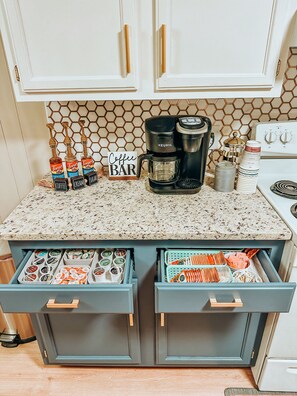 A kitchen with a coffee station, including a coffee maker, Torani syrups, and a sign that says "Coffee Bar". There are drawers with various food items and a hexagonal patterned backsplash.