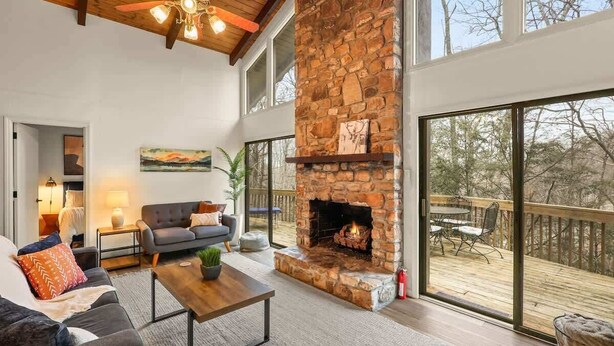 A living room with a stone fireplace, a ceiling fan, and large windows. - 4BR Chalet-A/C-New deck! Calm Creek-Quiet-2 Kings (Beech Mountain)