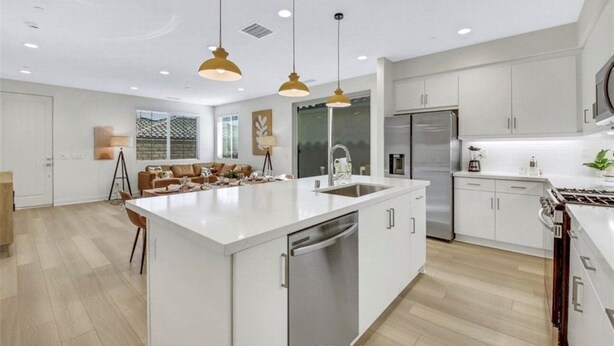 A modern kitchen with white cabinetry, a central island with a dishwasher, stainless steel appliances, pendant lighting, and a seating area with a sofa and coffee table. - PGA West-Catching Sunsets-3bd 2ba in Signature (La Quinta)