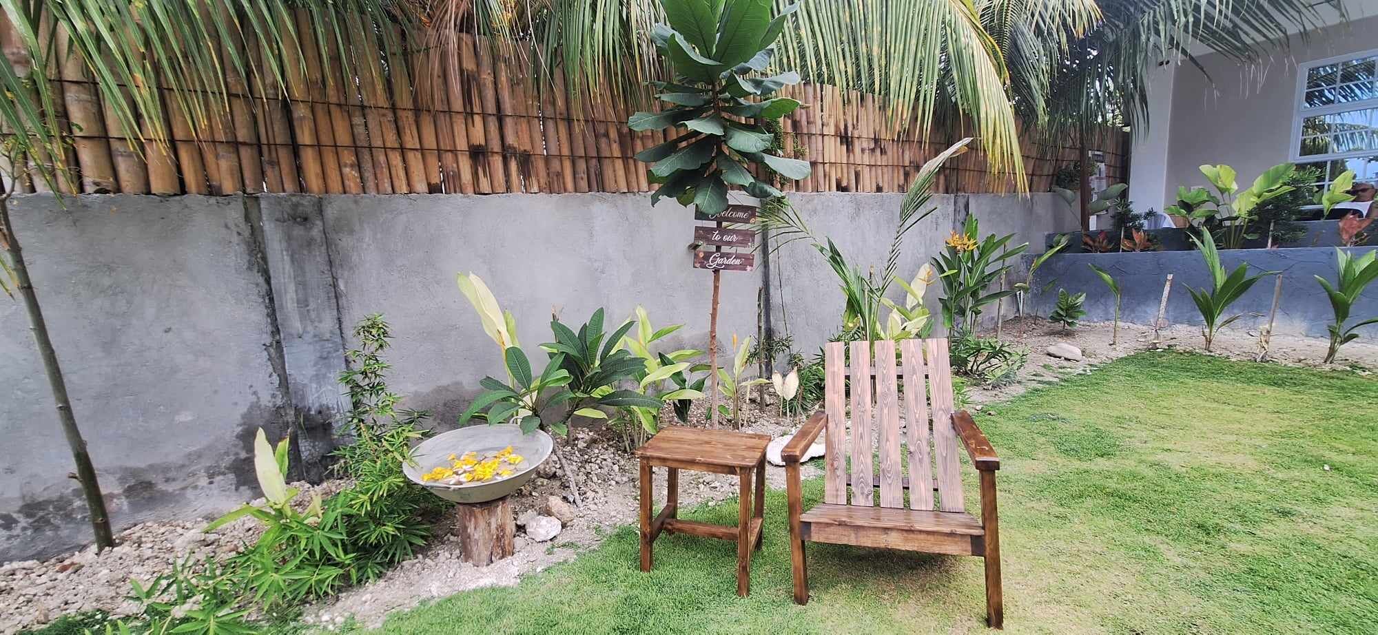 Un banco y una mesa de madera en un jardín con valla de bambú y diversas plantas.