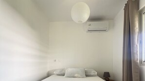 Een eenpersoonsbed met een witte spreideken, een bedlampje en een airconditioner aan de muur.