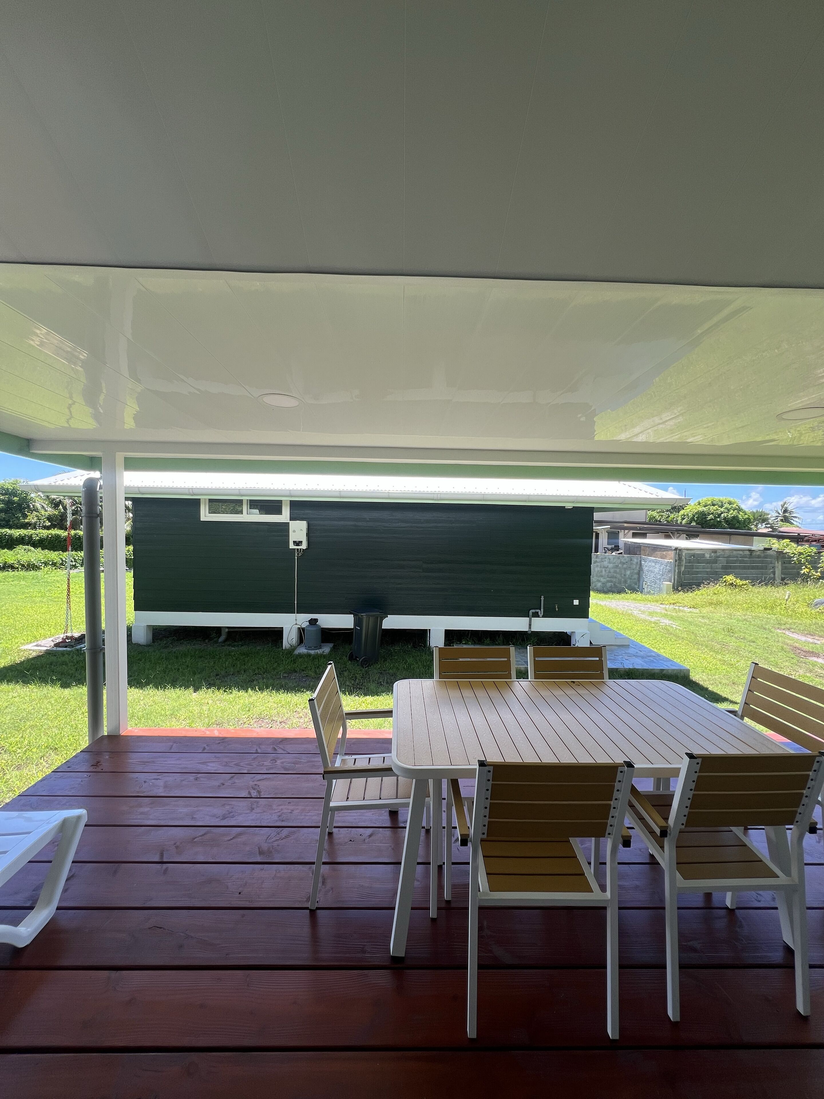 An outdoor patio with a wooden table and chairs, a covered area, and a green building in the background.