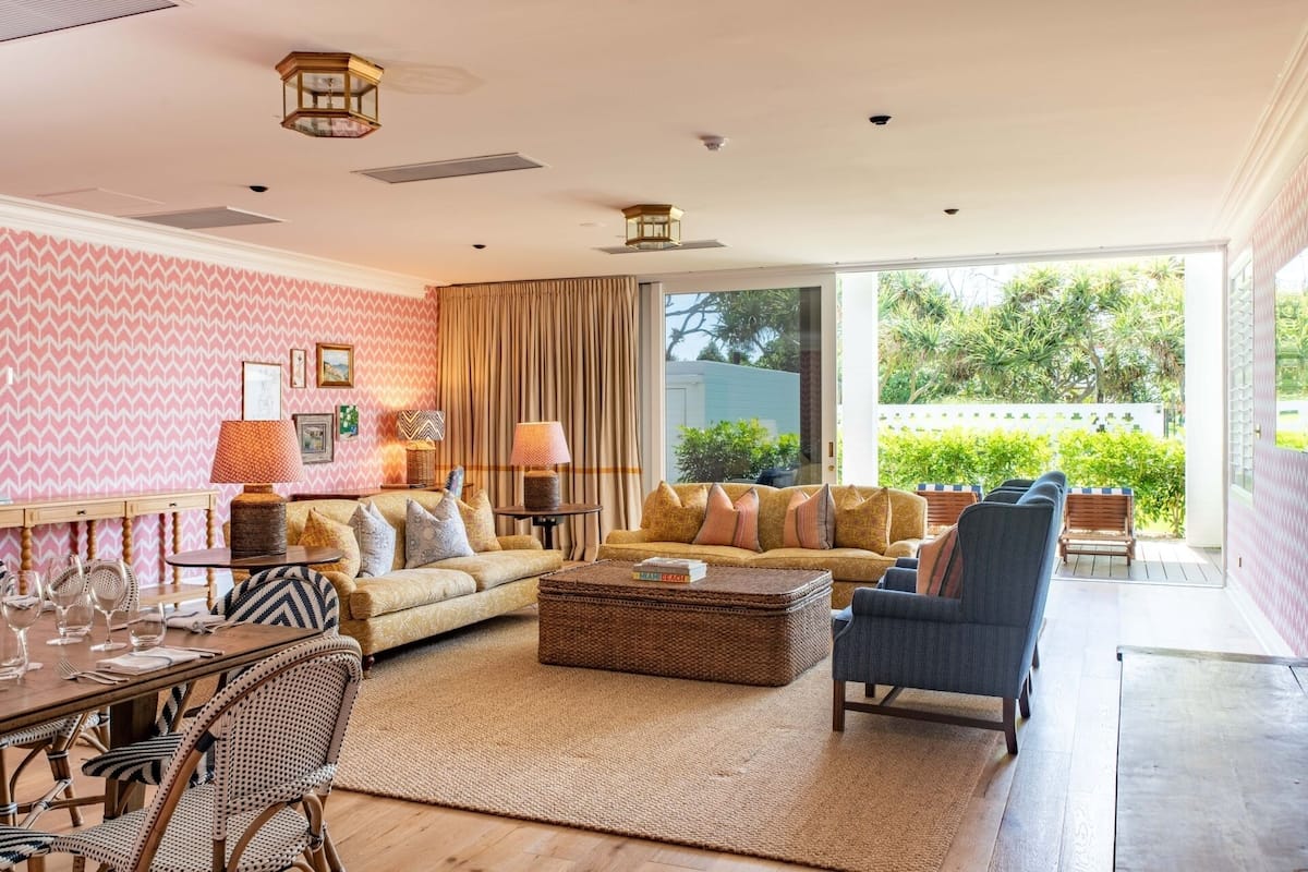 A spacious living room with a large sofa, a coffee table, and a dining area with wicker chairs. There are two lamps on side tables, and a view of greenery outside.