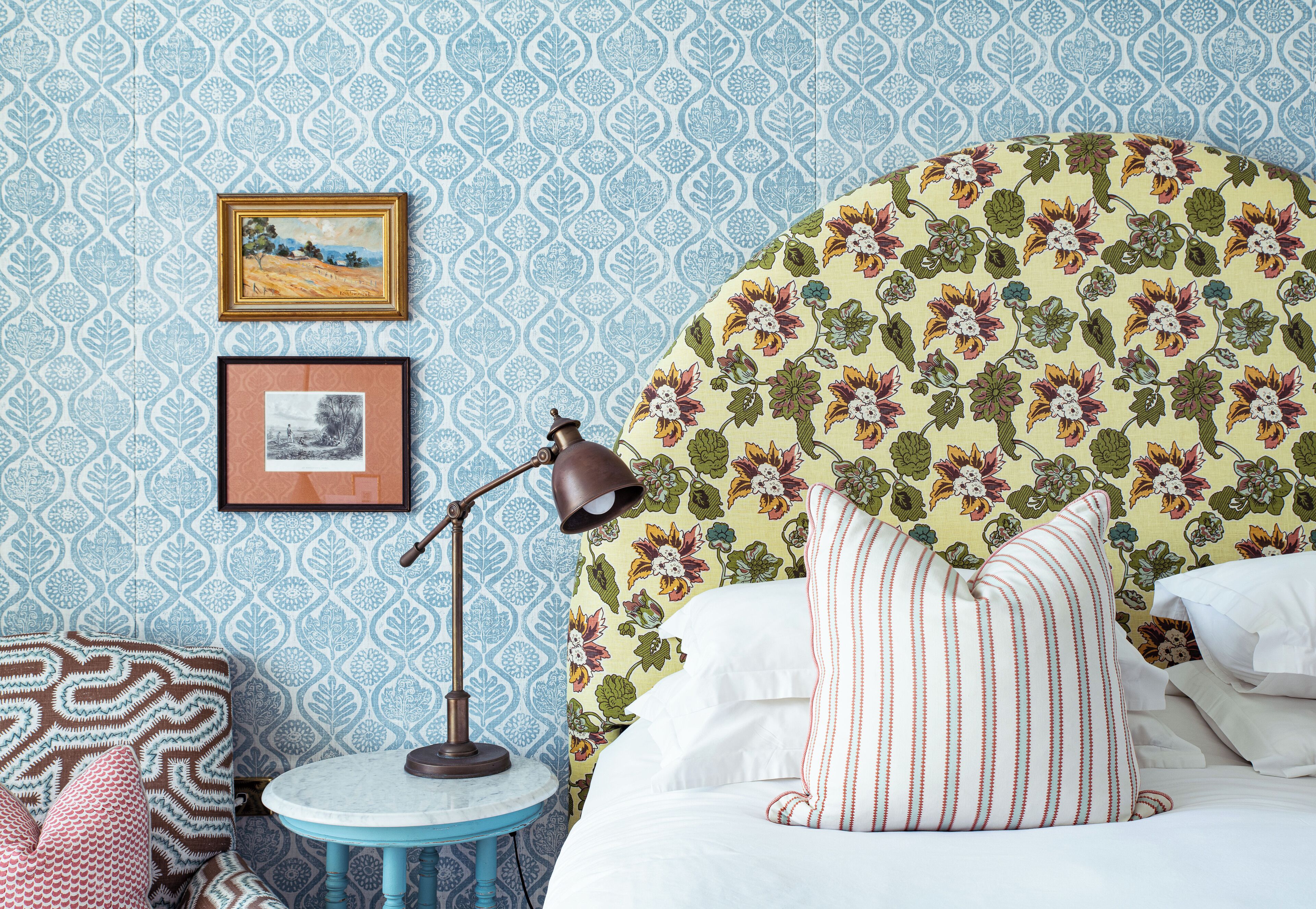 A bedroom with a patterned wallpaper, a round side table with a lamp, and a bed with floral headboard and striped pillows.
