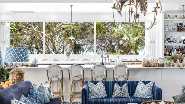 A modern living room with a blue sofa, wicker chairs, and a large window with a view of trees. - Halcyon House (Cabarita Beach)