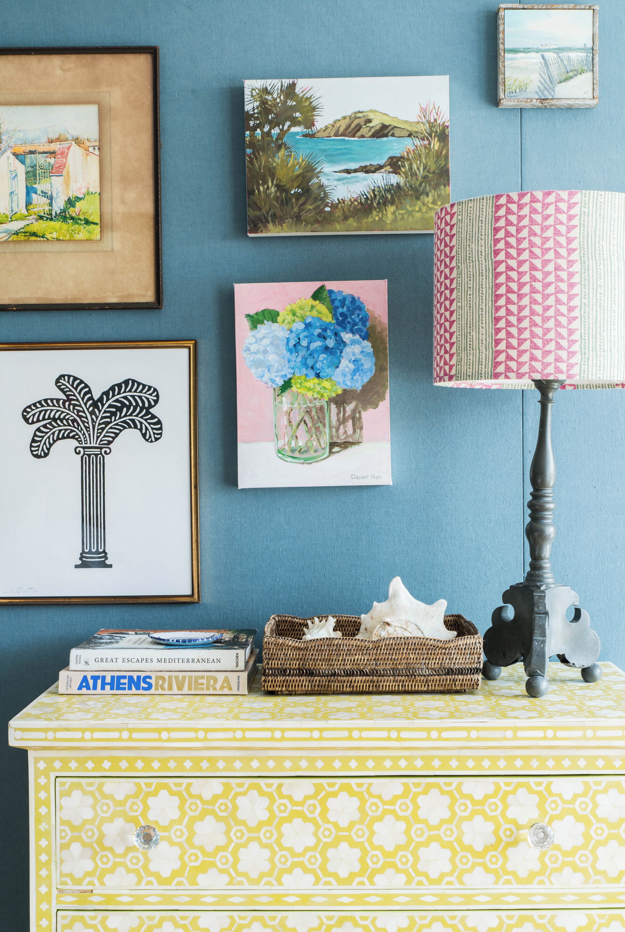 A wall with framed artwork, a lamp, and a small table with books and a basket.
