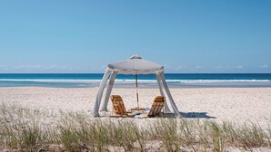 A beach umbrella with two wooden chairs under it, positioned on a sandy beach with the ocean in the background. - Halcyon House (Cabarita Beach)