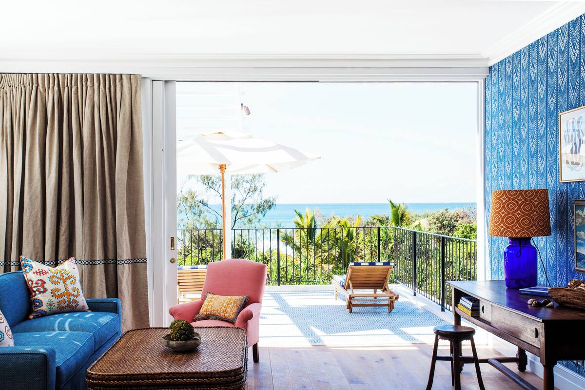 A room with a view of the ocean, a blue sofa, a pink armchair, and a wooden table with a lamp.