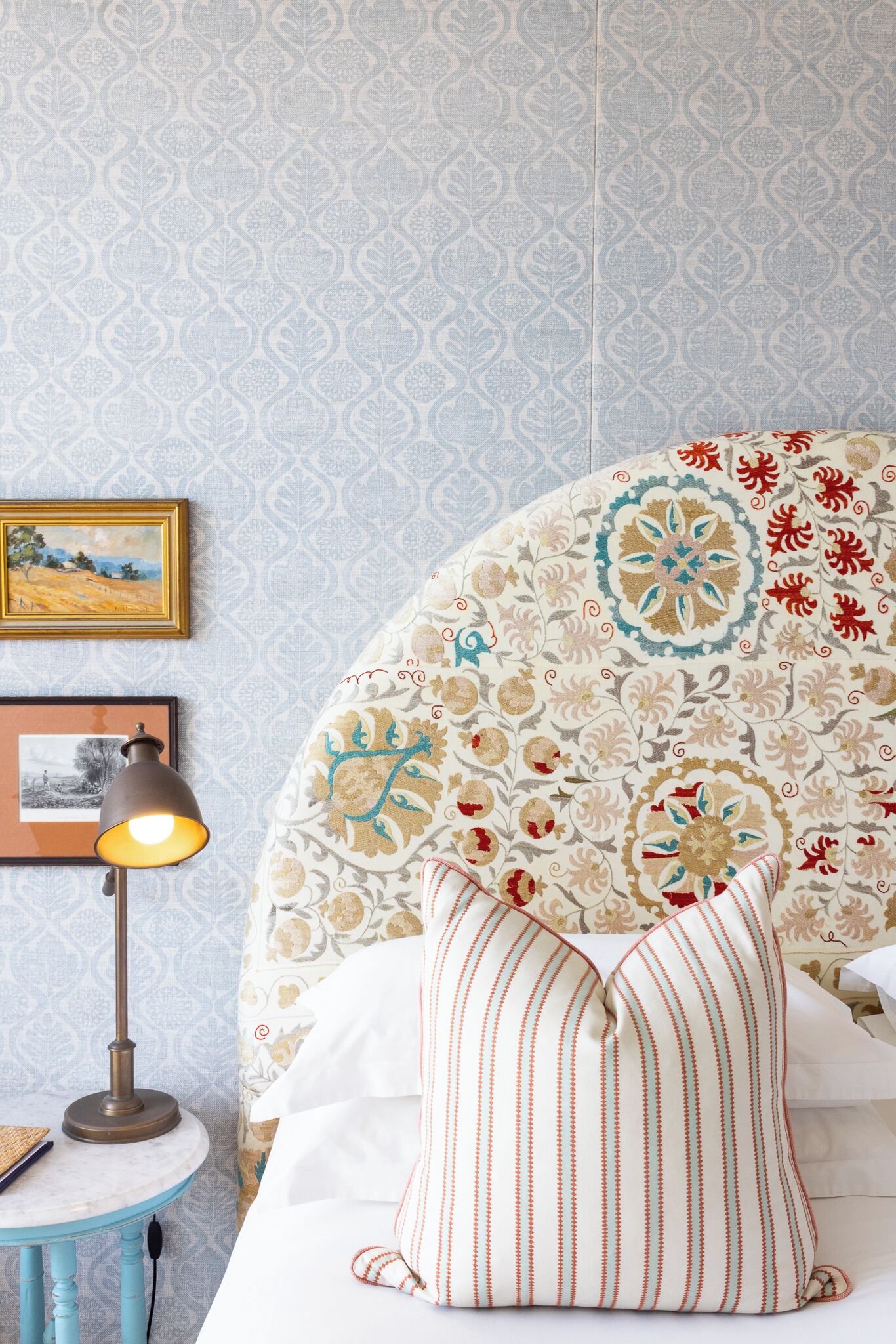 A bedroom with a patterned wallpaper, a round table with a lamp, and a bed with a decorative headboard and striped pillow.