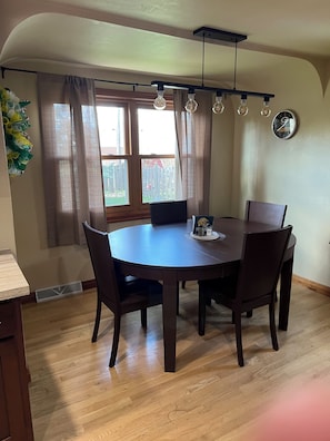 Un coin repas comprenant une table ronde, quatre chaises, une horloge au mur et une fenêtre avec des rideaux.