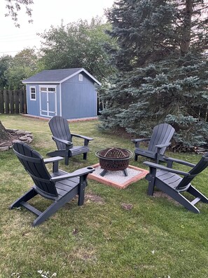 Un patio trasero con un brasero, cuatro sillas tipo Adirondack y un cobertizo.