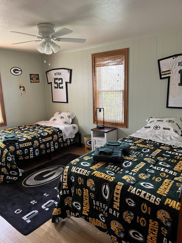 Une chambre avec deux lits, chacun agrémenté de linge de lit aux couleurs des Green Bay Packers, un ventilateur de plafond et une fenêtre avec des stores.